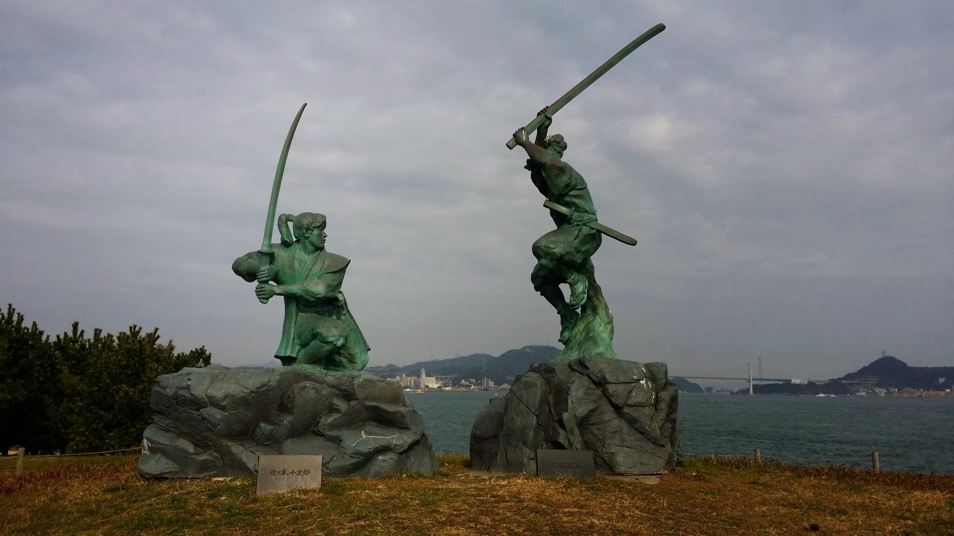 巌流島に立つ宮本武蔵・佐々木小次郎の決闘像。奥に関門海峡と関門橋が見える