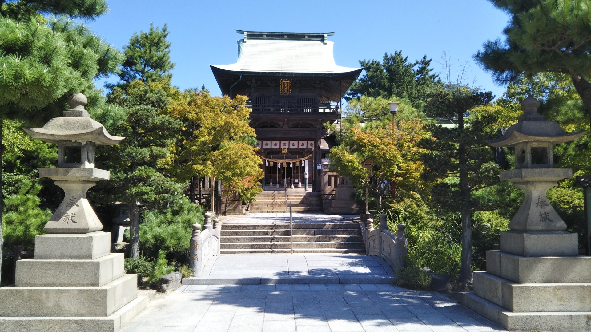 青空の下、石灯籠と参道の先にそびえる彦島八幡宮の楼門